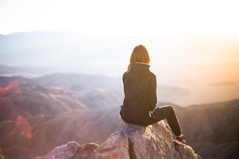 高い岩山の頂上に腰かけて朝焼けを見る女性の後ろ姿